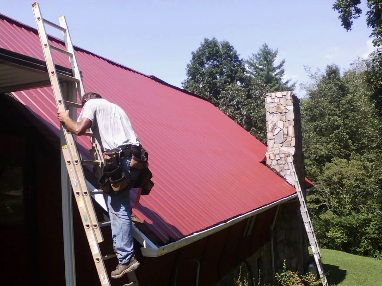 Roofer on a ladder beside a red metal roof and stone chimney — Soffit & Fascia Repair by Roofing Brothers in Nelsonville